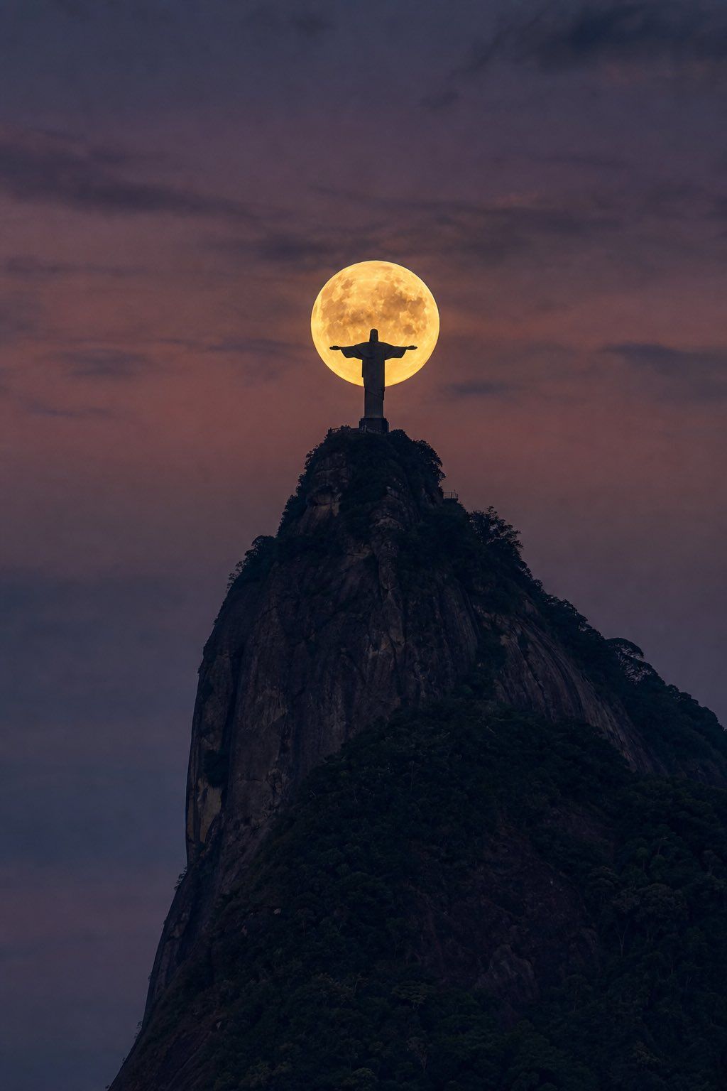 Christ the Redeemer silhouetted before a golden full moon atop Corcovado Mountain at purple dusk